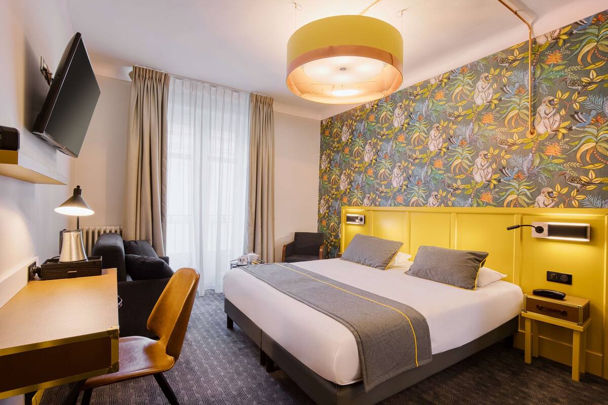 A hotel room with a bed, a desk, a chair, a TV, a lamp, and a patterned wall.