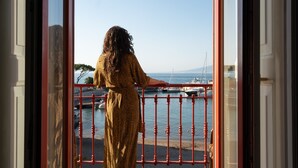 Una dona amb un vestit estampat, dempeus a un balcó amb vistes al mar i a vaixells.