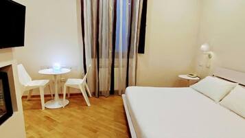 A hotel room with a bed, a small table with chairs, a TV mounted on the wall, and a window with curtains.