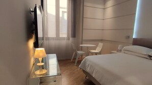 Habitación de hotel moderna con una cama grande, un escritorio pequeño con lámpara, una silla y una ventana con cortinas.
