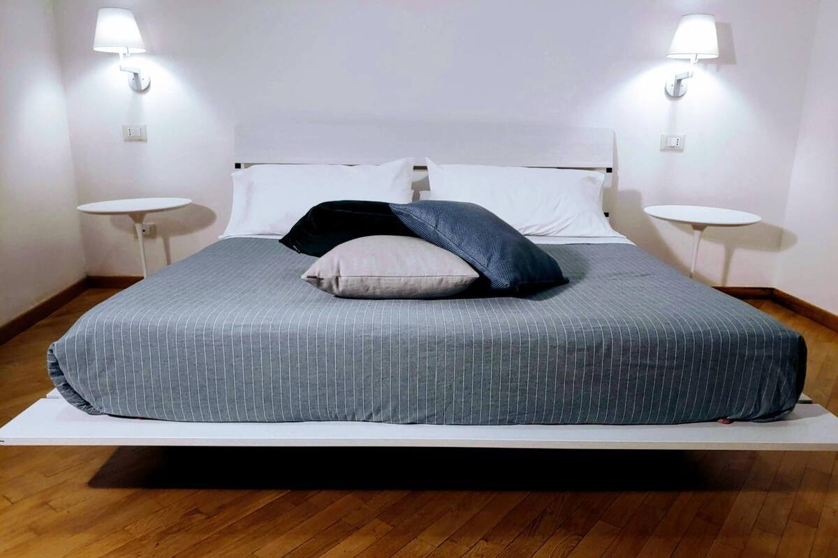 A neatly made bed with a grey striped bedspread, two white pillows, and a black pillow on a white mattress. There are two bedside tables with lamps on either side of the bed.
