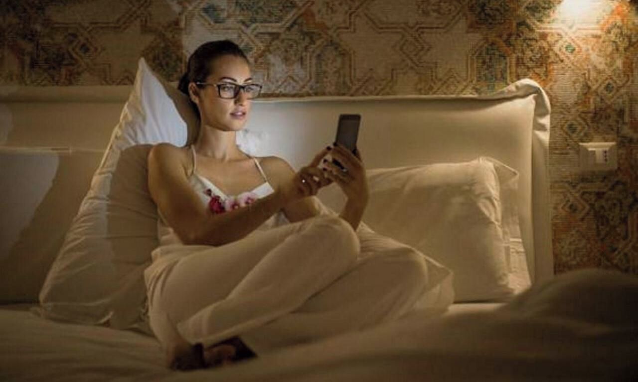 Eine Person sitzt auf einem Bett mit einem Kissen und hält ein Smartphone.
