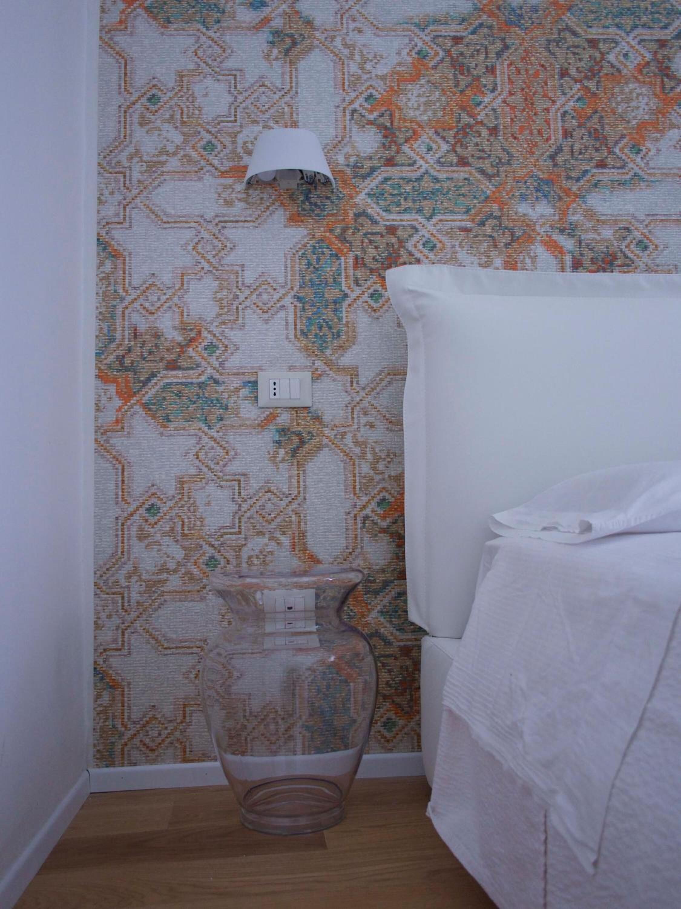 Une salle de bain avec un papier peint à motifs, des toilettes blanches, un vase en verre transparent et une serviette blanche posée sur le sol.