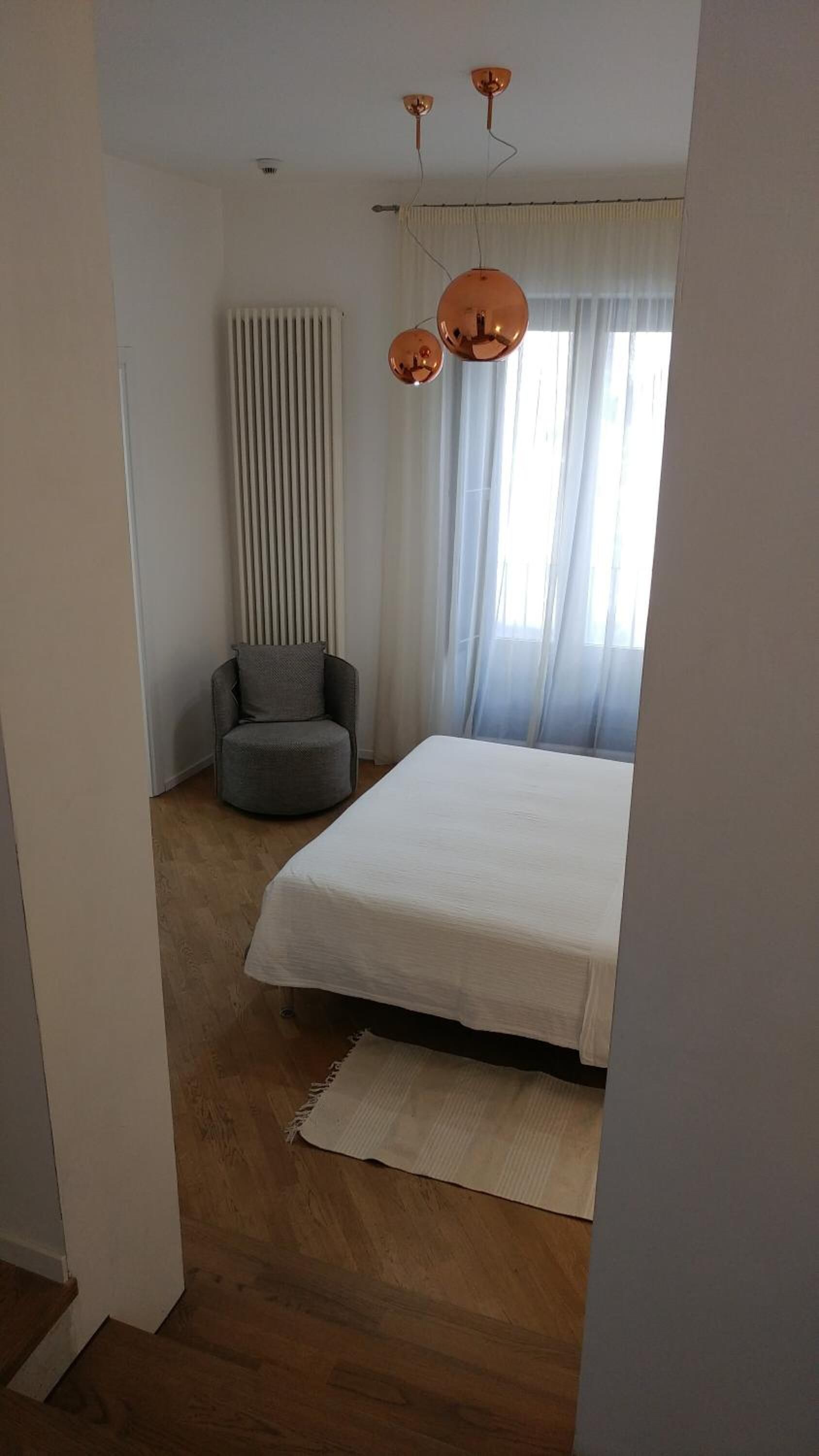 Un dormitorio con cama, una silla, un radiador y una ventana con cortinas.