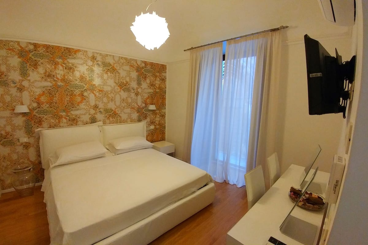 A bedroom with a bed, a desk, a chair, a TV, and a window with curtains.