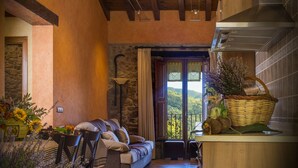 A cozy living area with a sofa, chairs, and a dining table. There is a kitchen area with a stove and a window overlooking greenery. - Rusticae La Casassa de Ribes (Ribes De Freser)