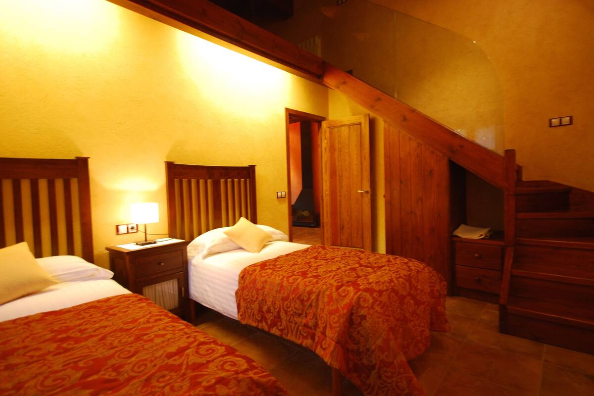 Habitación de hotel con dos camas, cabeceros de madera, una mesita de noche con lámpara y una escalera de madera.