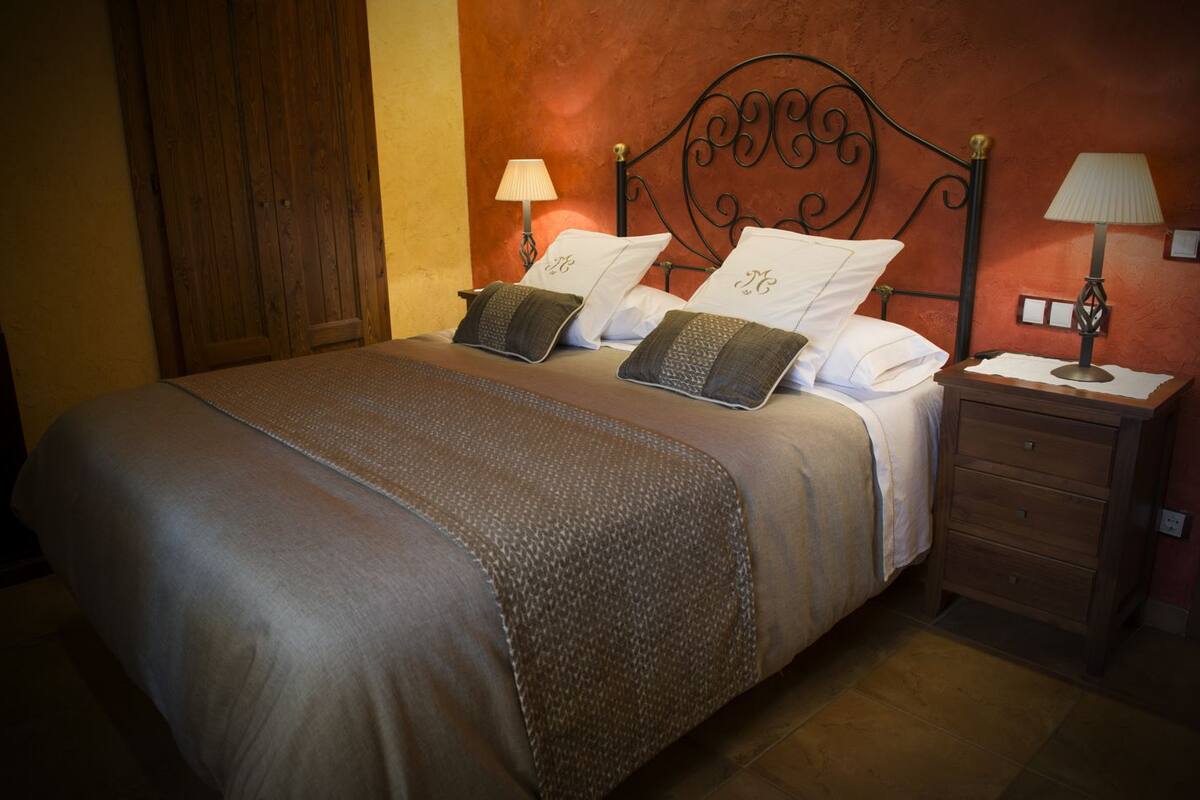 Una cama bien tendida con una colcha estampada, dos almohadas con iniciales bordadas, un cabecero de metal, lámparas de noche y una mesita de noche de madera.