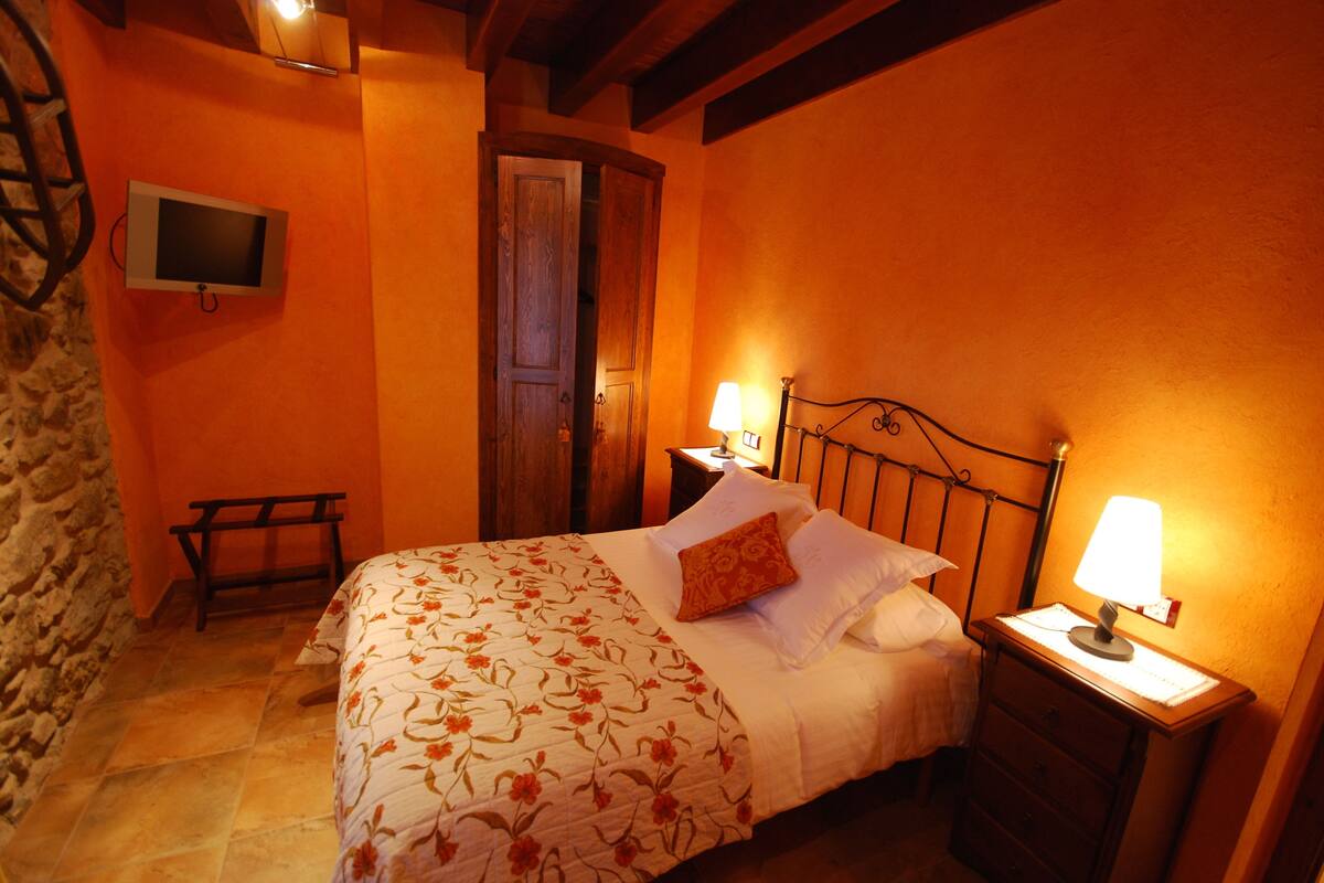 Un dormitorio con cama, mesitas de noche, un televisor montado en la pared y una puerta de madera.