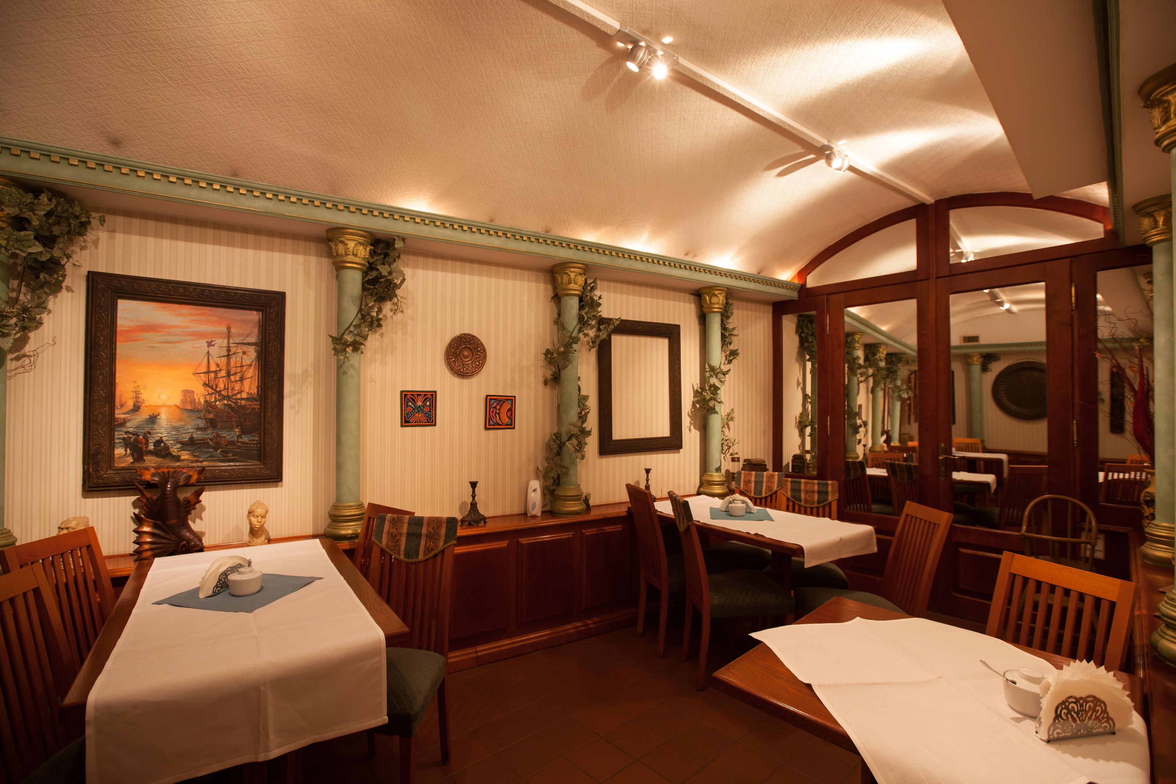 Un comedor con mesas dispuestas para el servicio, sillas de madera y una pared decorativa con obras de arte enmarcadas y plantas.