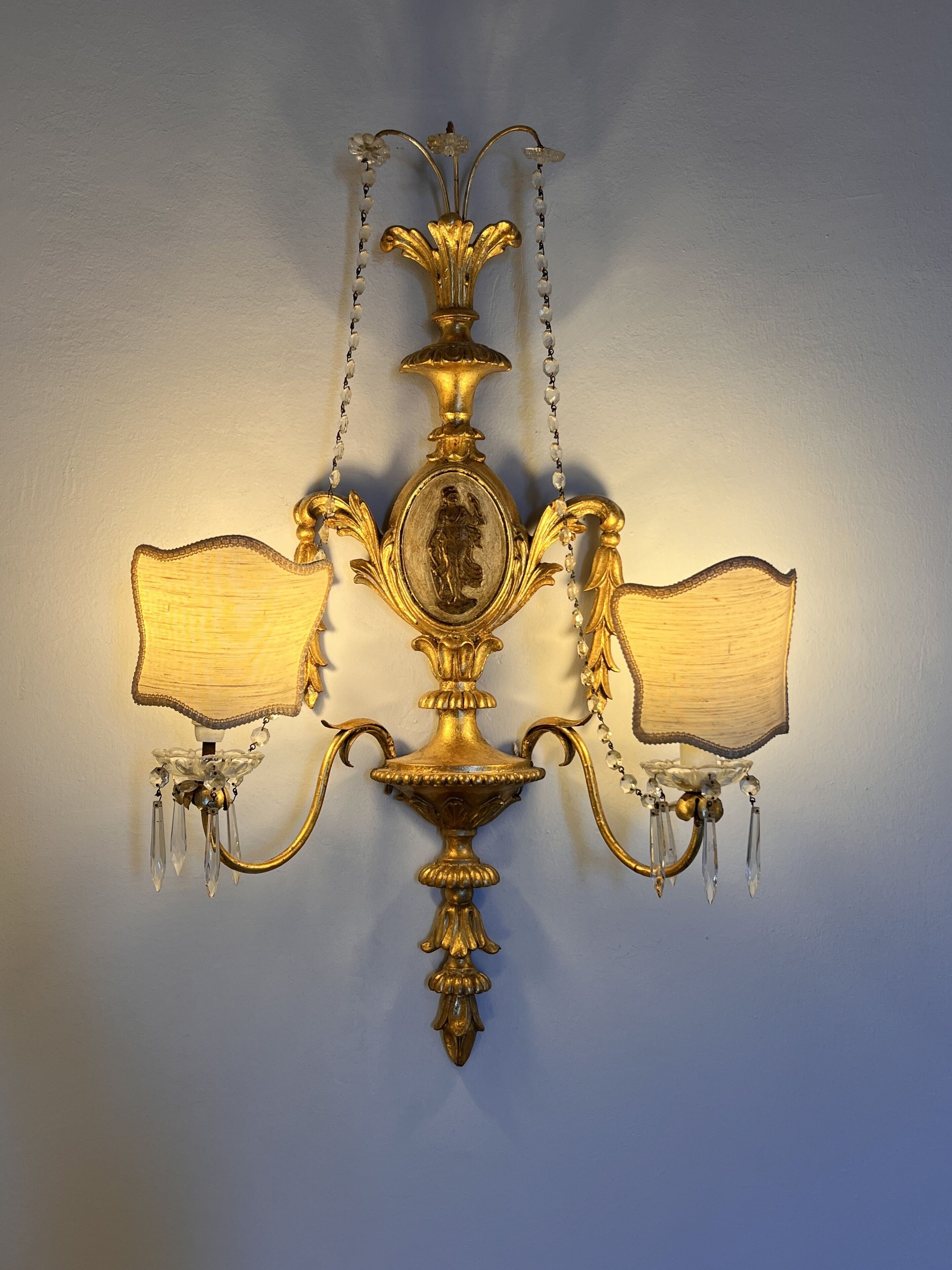 A wall-mounted chandelier with a central ornate design and two lampshades on either side.