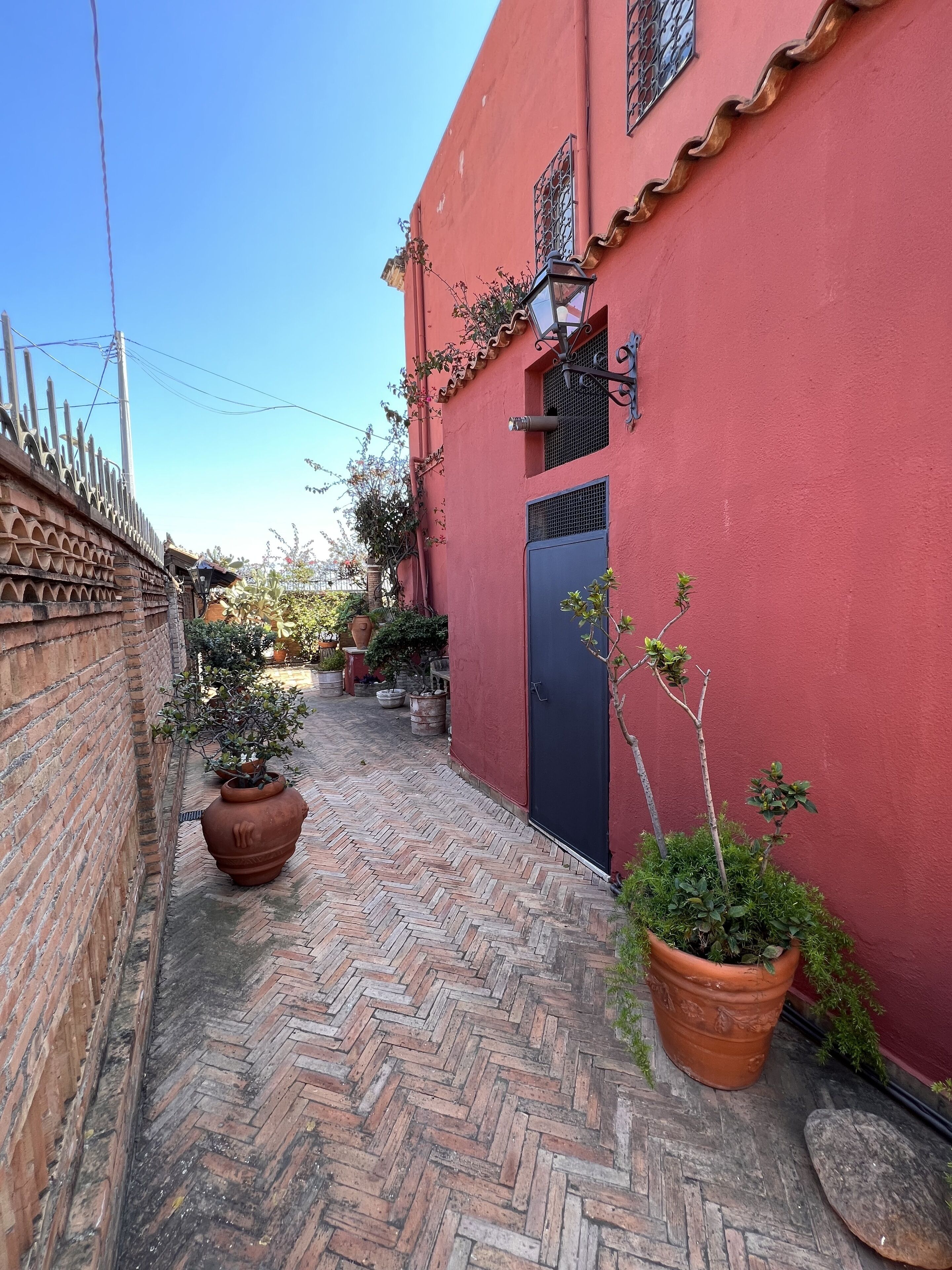 Une ruelle étroite avec un bâtiment rouge, des plantes en pot et un sol en briques disposées en motif chevrons.