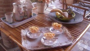 Una mesa con una bandeja de postres, una jarrón, tazas y un cuenco de fruta.