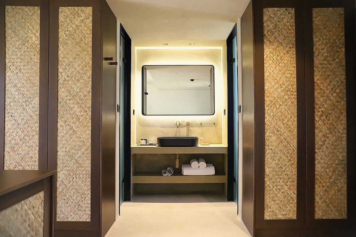 A narrow hallway leading to a bathroom with a large mirror, a sink, and a shelf with towels.