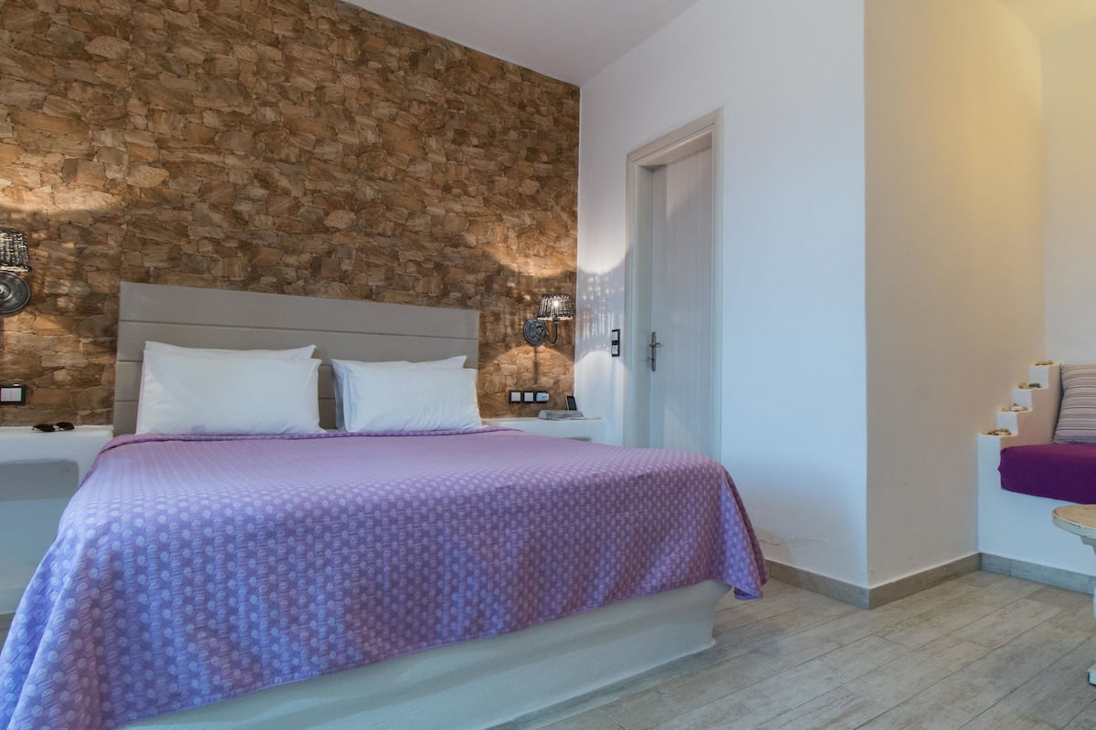 A bedroom with a stone wall, a bed with a purple bedspread, a nightstand, a lamp, and a wooden table with fruits.