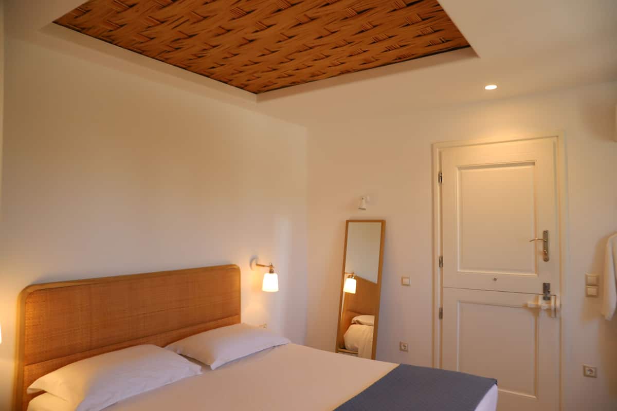 A hotel room with a wooden ceiling, a bed with white linens, a nightstand with a lamp, and a door with a towel hanging on it.