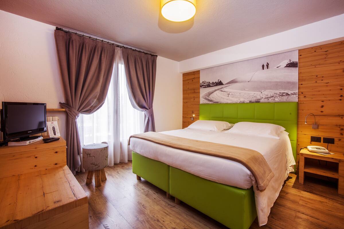 A bedroom with a green upholstered bed, a wooden nightstand, a television on a wooden dresser, and a painting on the wall.