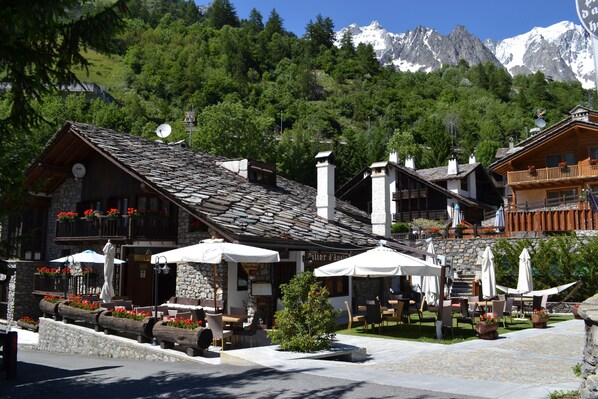 A traditional chalet-style building with stone walls and a sloped roof, outdoor seating with umbrellas, and a view of snow-capped mountains in the background. - Pilier d'Angle (Courmayeur)