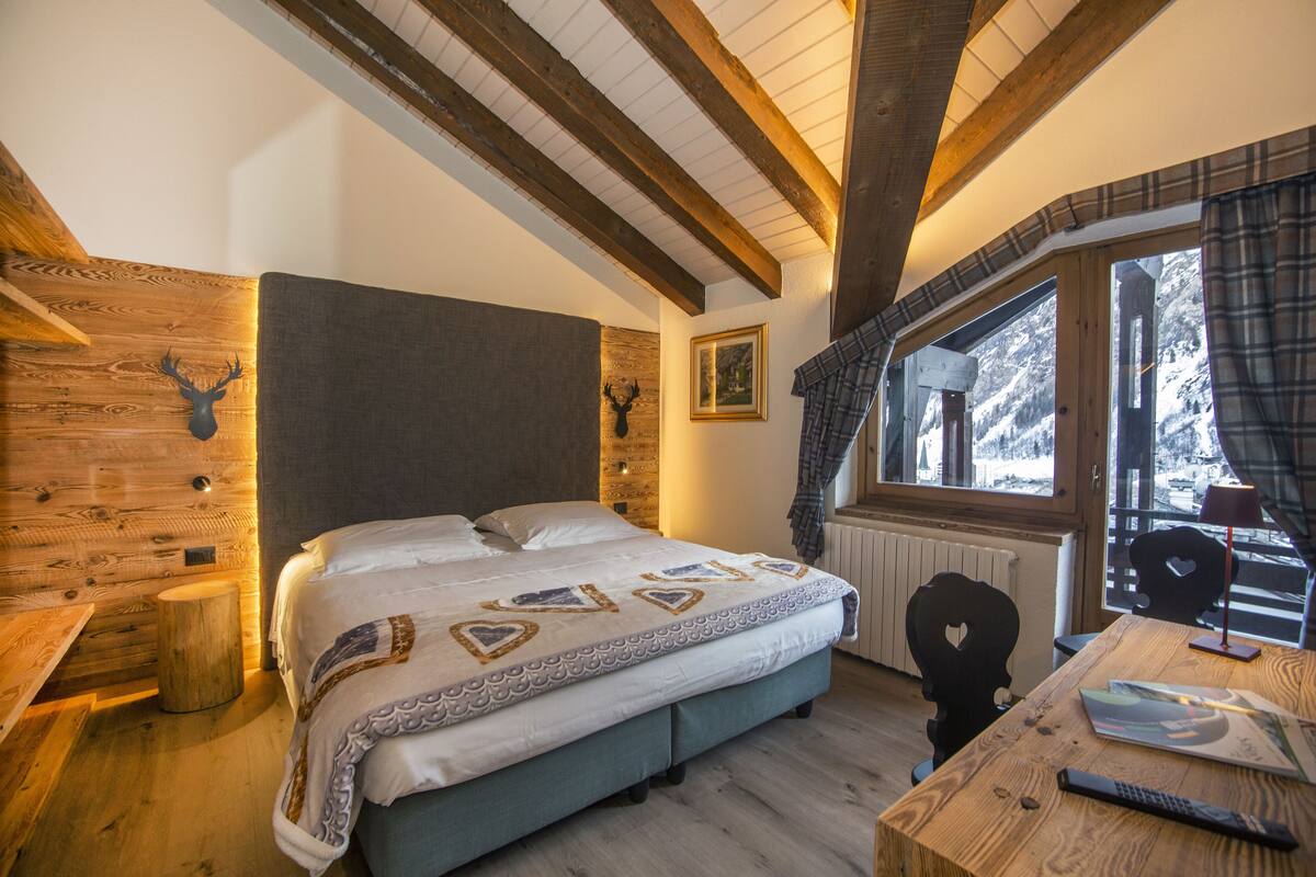 A cozy room with a bed, a wooden headboard, a window with plaid curtains, and a table with a lamp.