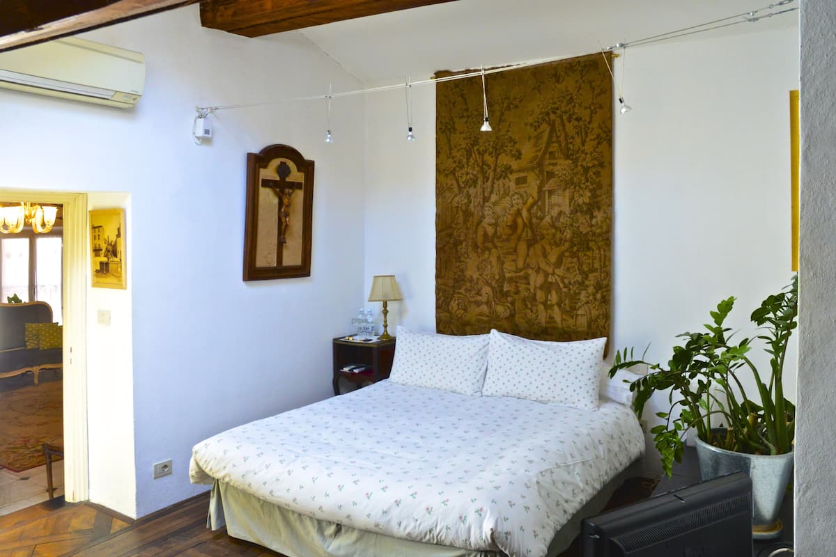 Een slaapkamer met een bed, nachtkastje, lamp, schilderij en een potplant.