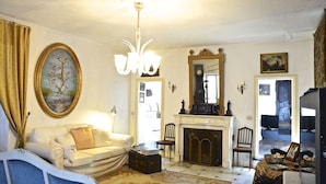 Een woonkamer met een witte bank, een open haard, een kroonluchter en een groot schilderij in een lijst.