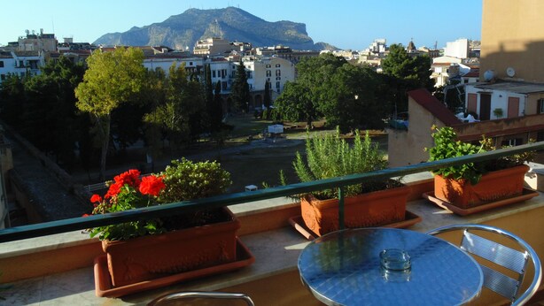 A balcony with potted plants, a table, and chairs, overlooking a cityscape with buildings and a mountain. - Bed and Breakfast Kemonia (Palermo)