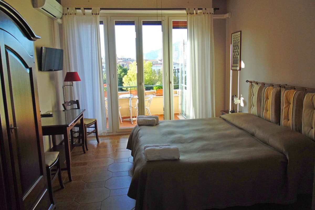 Habitación de hotel con cama, escritorio con lámpara, una silla y vistas al exterior por un ventanal corredero.