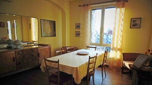 A dining area with a table set for four, wooden chairs, a window with curtains, and a view of buildings outside. - Bed and Breakfast Kemonia (Palermo)