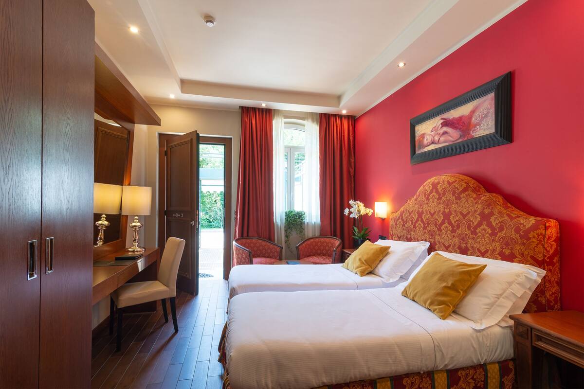 A hotel room with two beds, a desk with a lamp, a chair, and a large window with curtains.