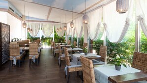 Un comedor interior con sillas de mimbre, manteles blancos, faroles colgantes y ventanas grandes con cortinas drapeadas.