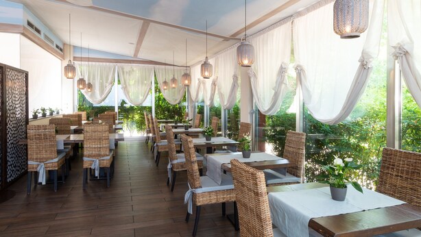 An indoor dining area with wicker chairs, white tablecloths, hanging lanterns, and large windows with draped curtains. - Hotel Silver (Milan)