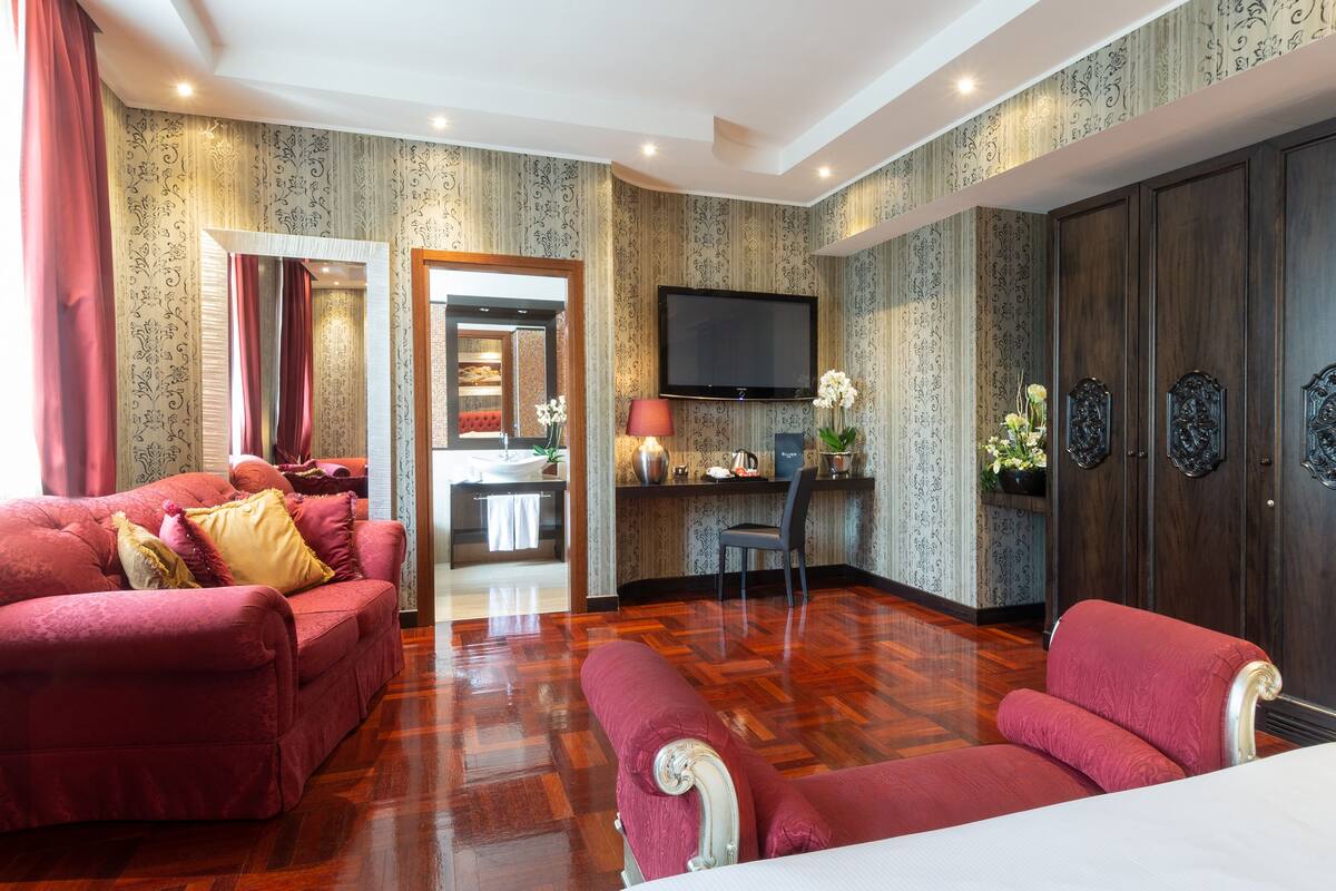 A hotel room with a red sofa, a matching armchair, a television, and a wooden wardrobe.