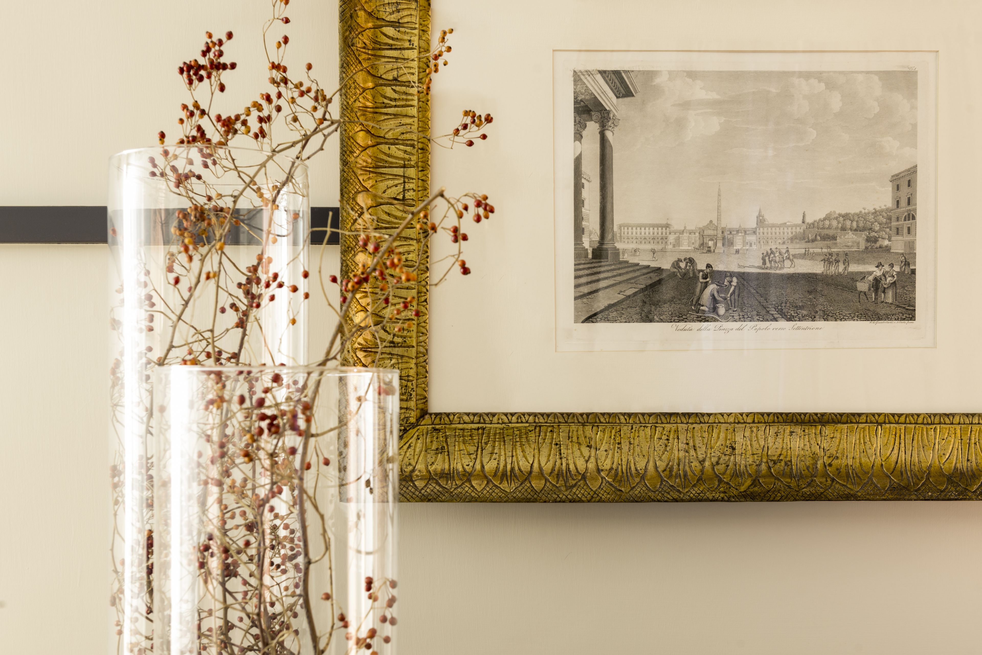 A framed black and white historical illustration on a wall, a clear vase with dried branches, and a decorative golden trim.