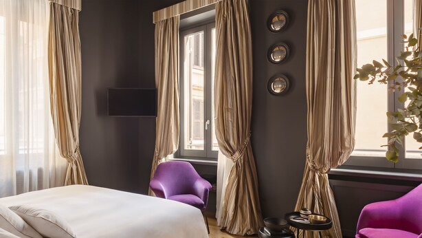 A hotel room with a bed, two purple chairs, a small table with a bowl, and curtains. - Crossing Condotti (Rome)