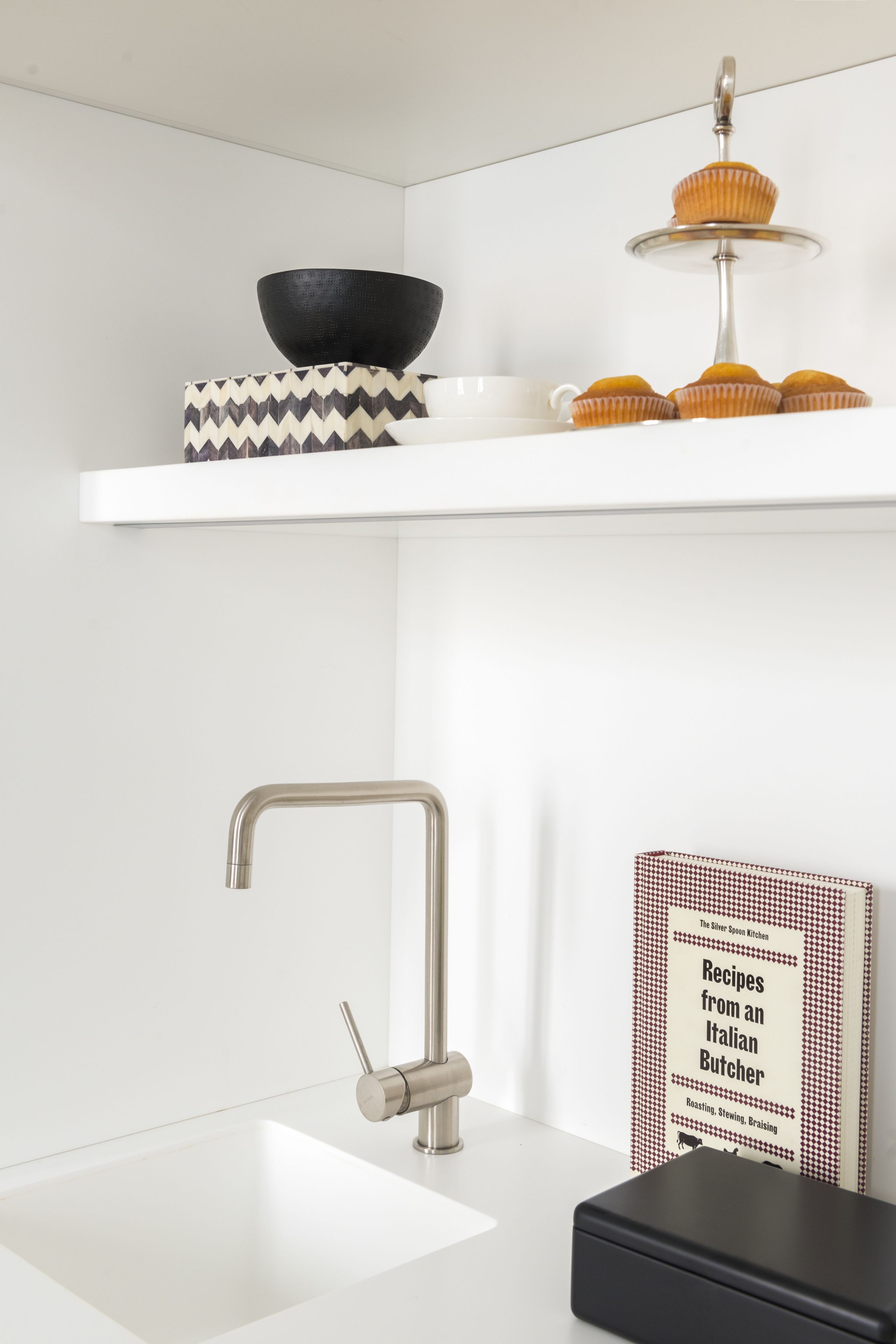 A modern kitchen with a white sink, stainless steel faucet, a black bowl, a patterned box, a book titled 'Recipes from an Italian Butcher', and a tiered stand with muffins.