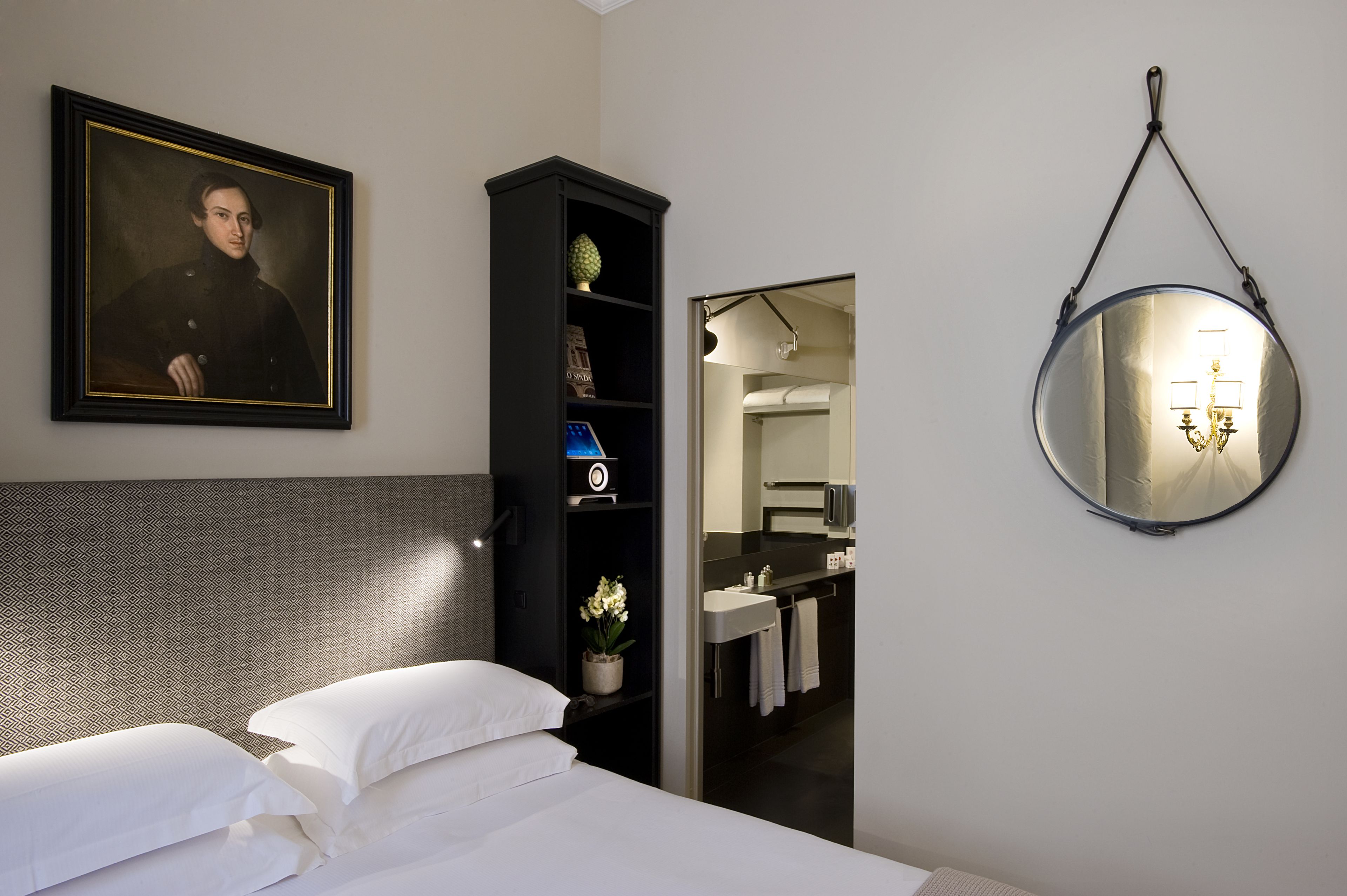 Une chambre d’hôtel avec un lit, un portrait encadré, un grand meuble noir et un miroir rond.