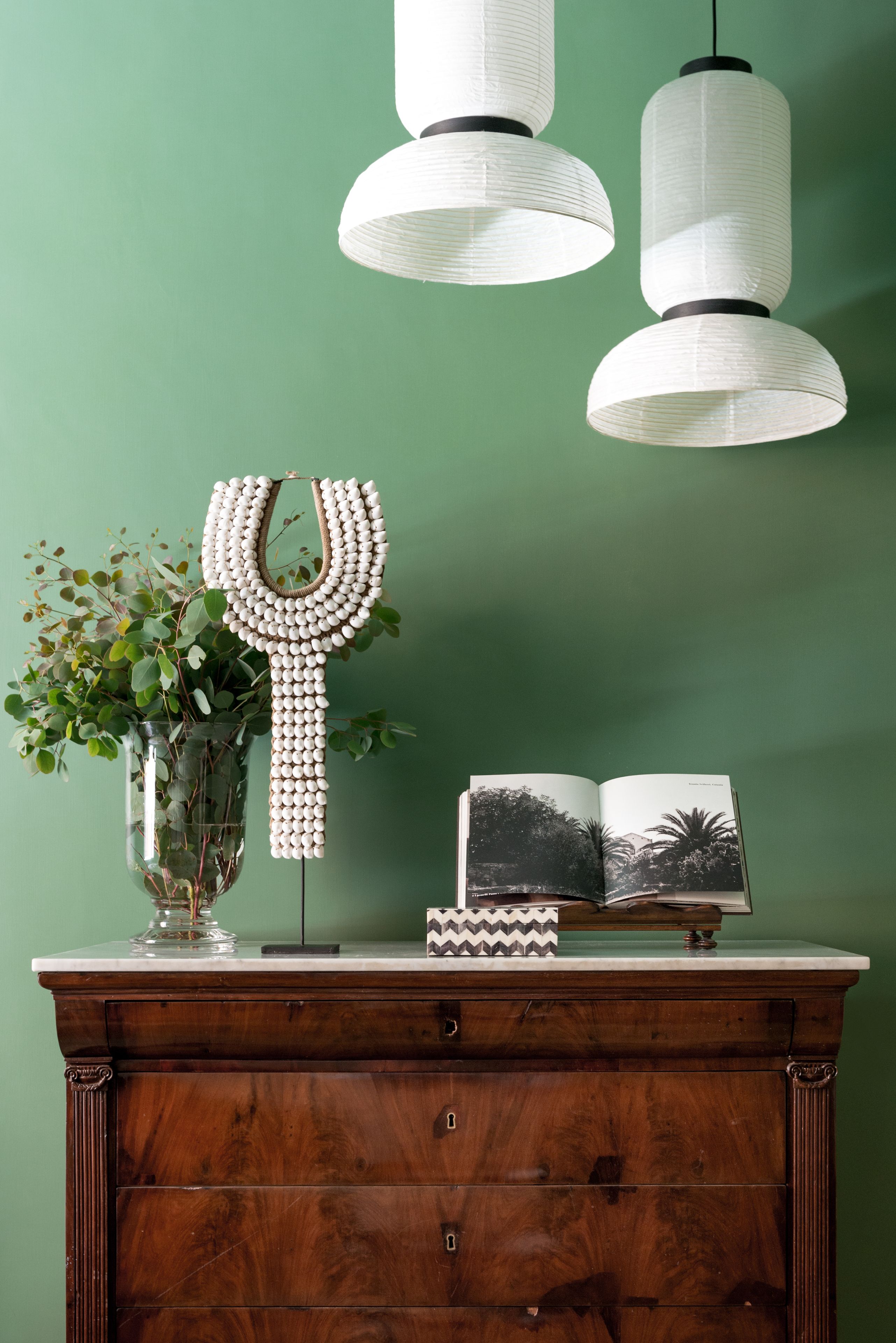 A wooden dresser with a clear glass vase containing green foliage, a decorative mirror, and an open book with a black and white photograph.