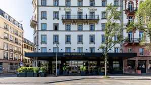 Un hôtel à plusieurs étages, de style architectural européen classique, qui se distingue par son entrée imposante, agrémentée d’un auvent et de plantes en pot.