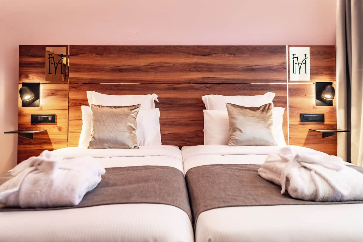Une chambre d’hôtel avec deux lits, chacun doté d’une tête de lit en bois, de draps blancs et d’une paire de serviettes. On y trouve des lampes fixées au mur et une petite étagère avec une lampe.