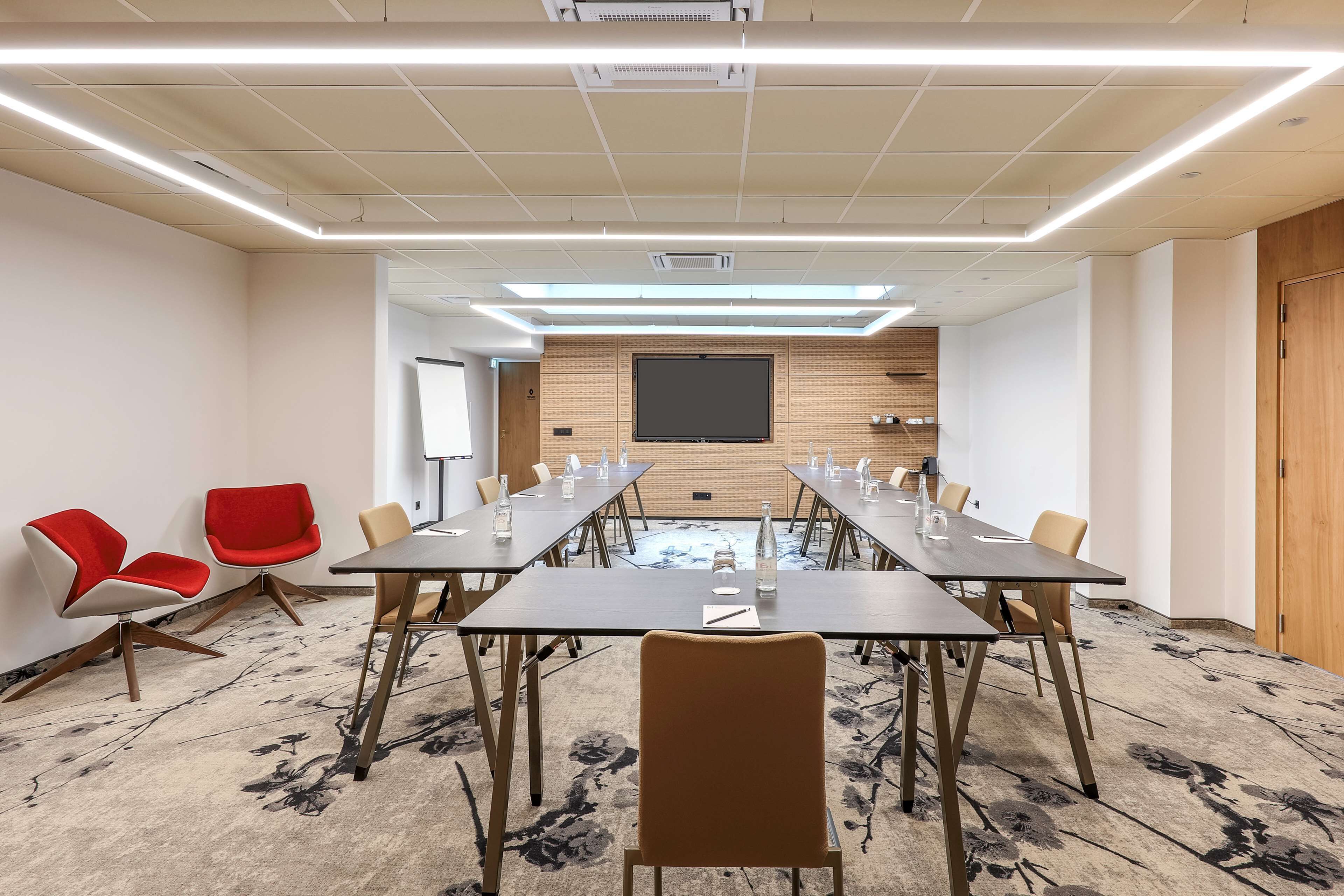 A conference room with a long table, red and beige chairs, a projector screen, and a wooden panel wall.