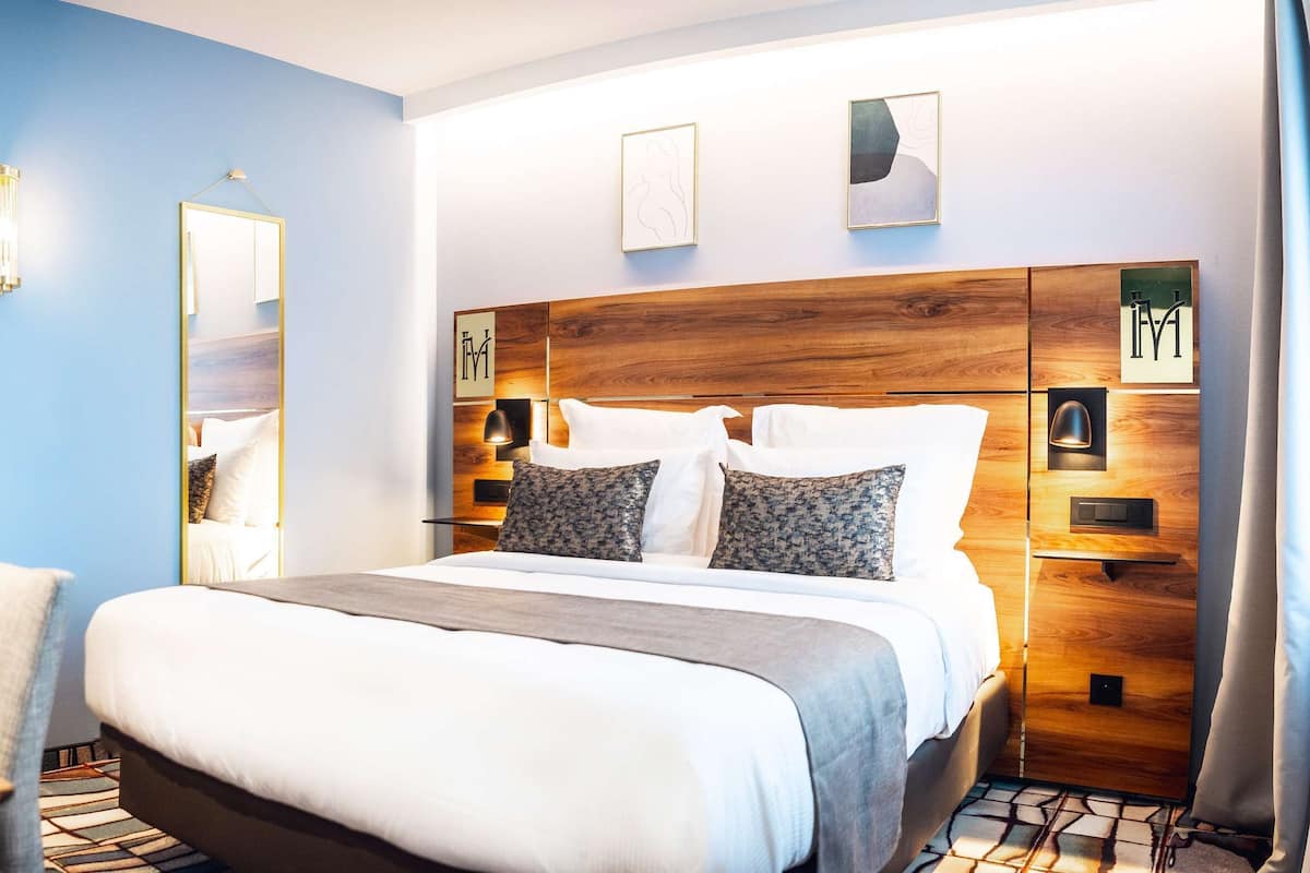 Une chambre d’hôtel comprenant un lit, une table de chevet, une lampe et une tête de lit en bois agrémentée d’une décoration murale.