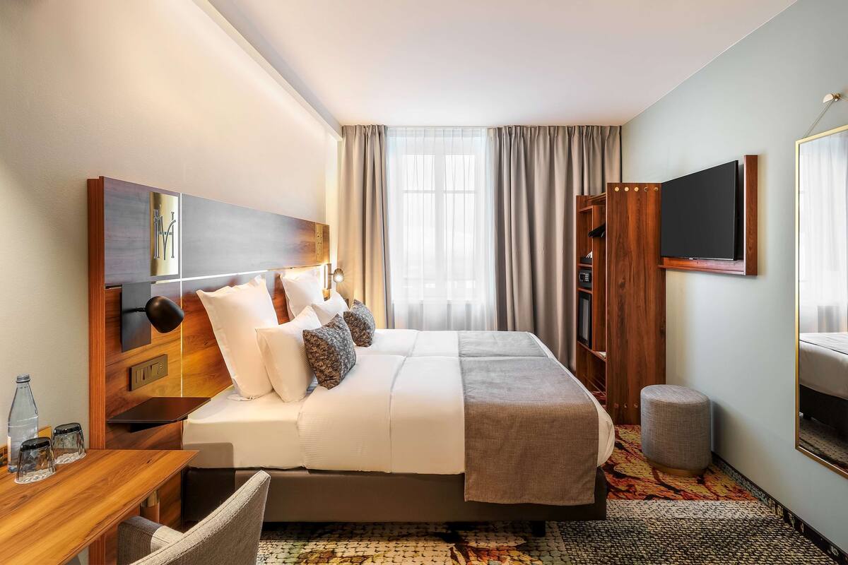 Une chambre d’hôtel avec un grand lit, un bureau avec une lampe, un téléviseur fixé au mur et une fenêtre avec des rideaux.