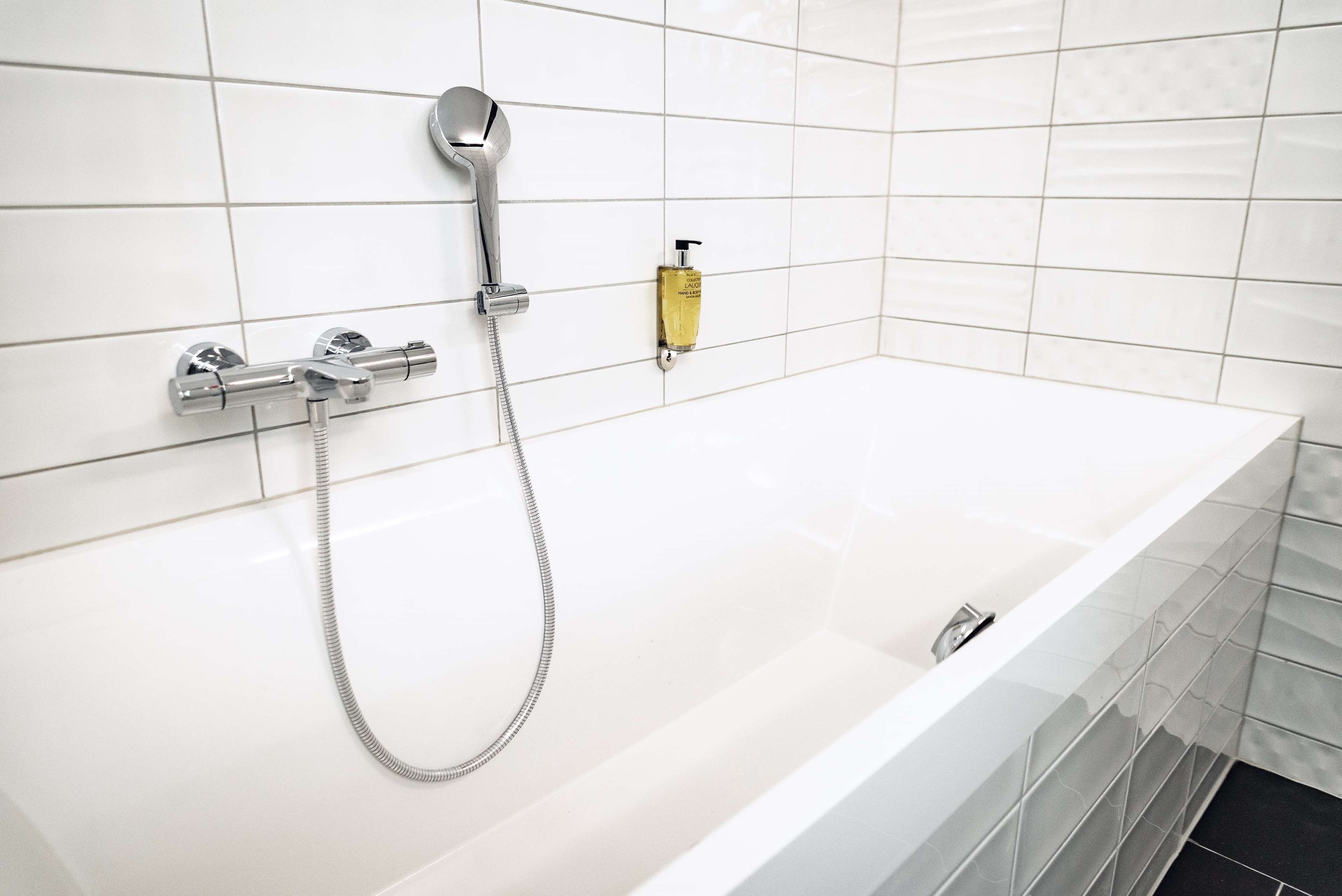 A modern bathroom with a white bathtub, a handheld showerhead, and a bottle of shampoo.
