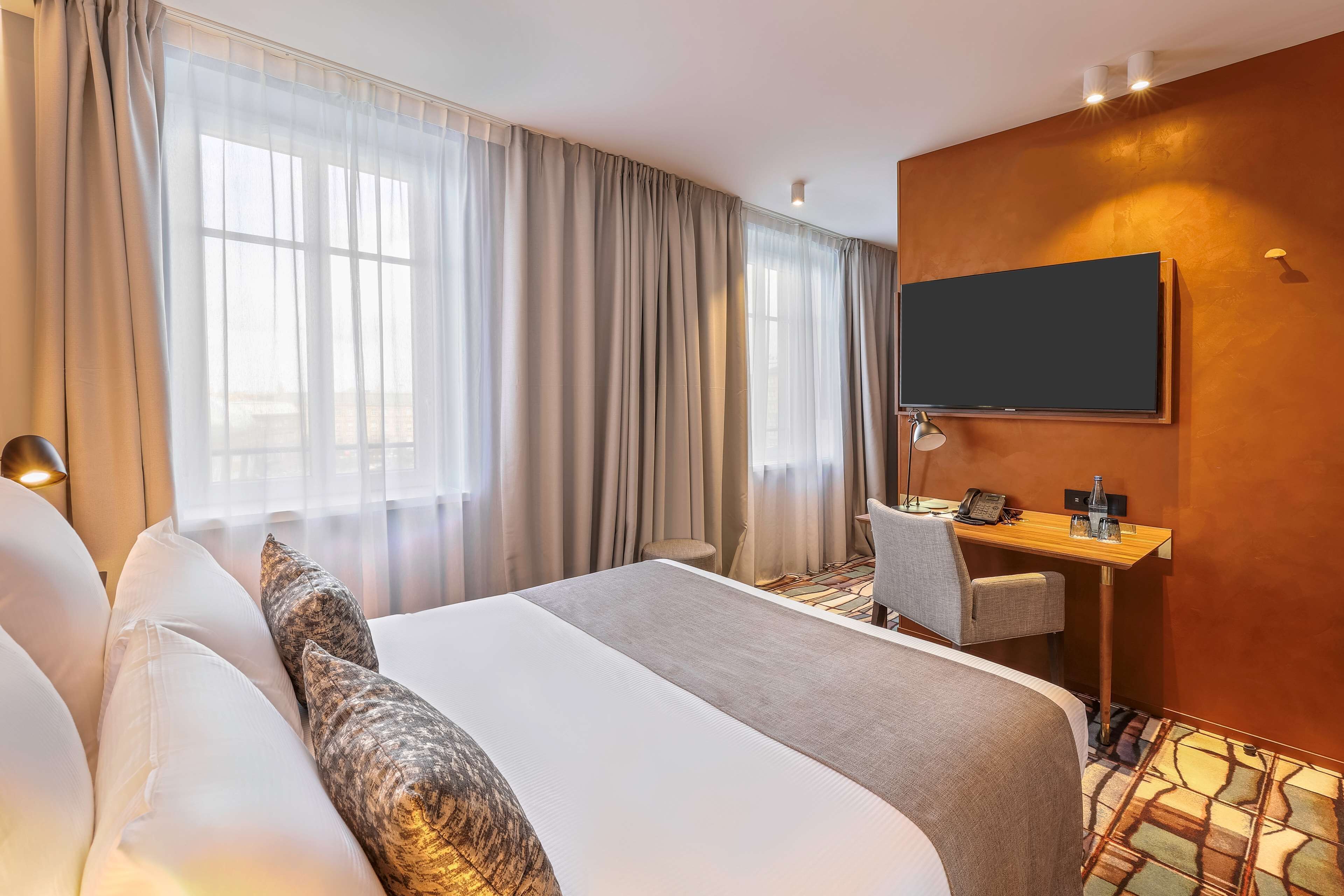 Ein Hotelzimmer mit einem großen Bett, einem Schreibtisch mit Stuhl, einem Flachbildfernseher und einem Fenster mit Vorhängen.