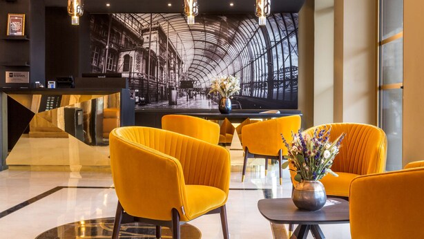 Un hall d’entrée avec un comptoir de réception, des chaises jaunes, un vase rempli de fleurs et une grande fresque représentant une gare.