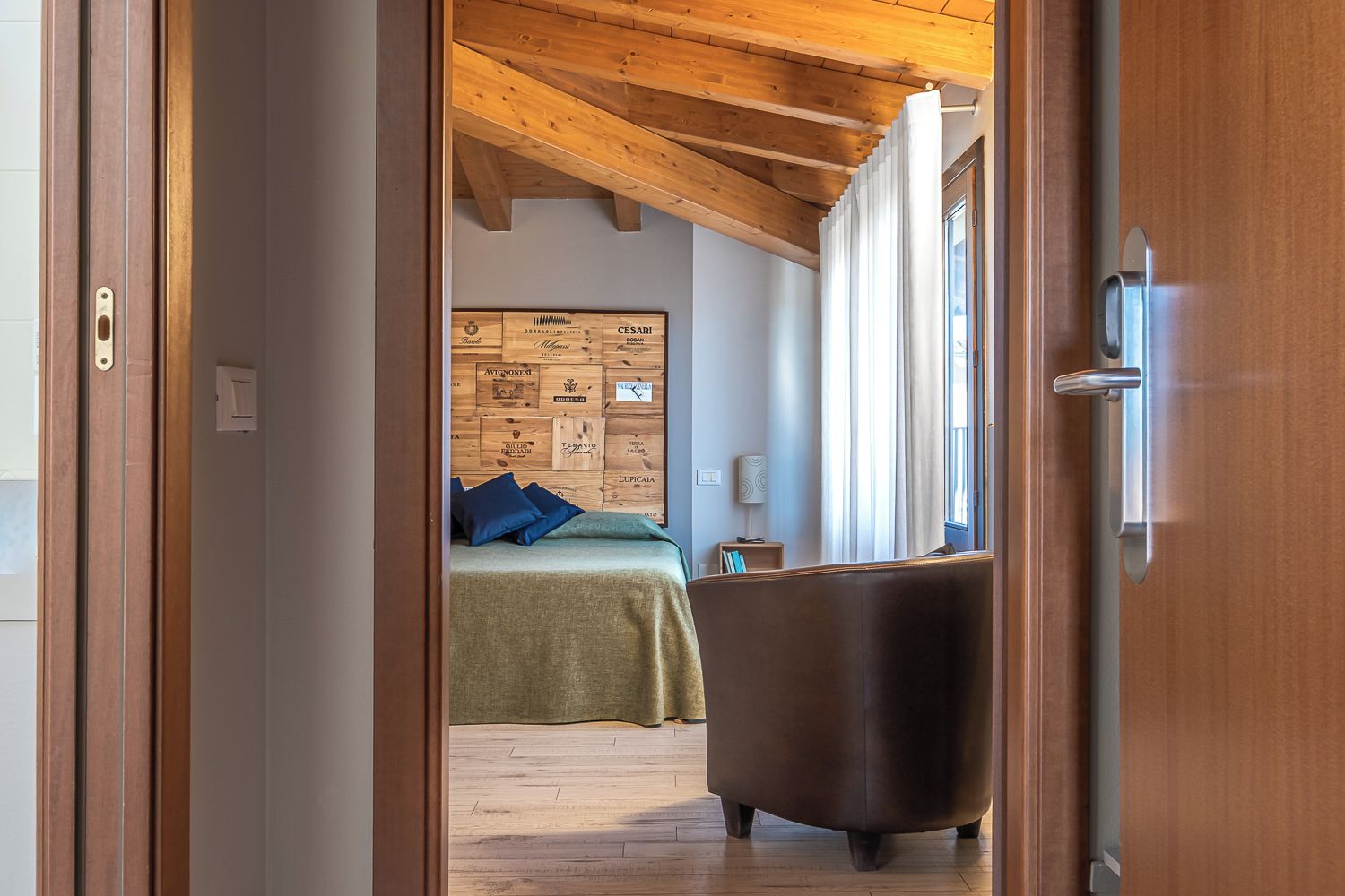 Una stanza con soffitto in legno, un letto con copriletto verde e cuscini blu, una poltrona scura e un pannello di legno con diverse etichette.