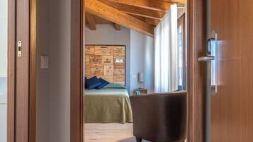 Una stanza con soffitto in legno, un letto con copriletto verde e cuscini blu, una poltrona scura e un pannello di legno con diverse etichette.