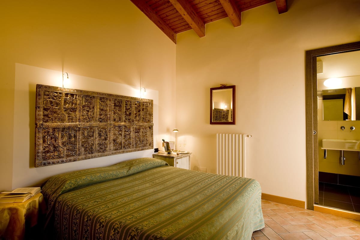 A bedroom with a large bed, a nightstand, a wall-mounted lamp, a radiator, and a bathroom door.