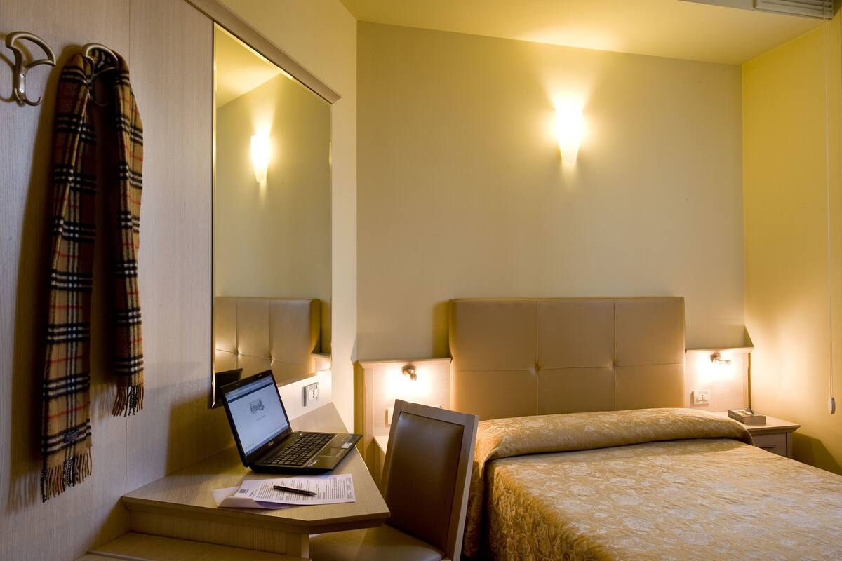 A hotel room with a bed, a desk with a laptop, a chair, a nightstand with a lamp, and a mirror.