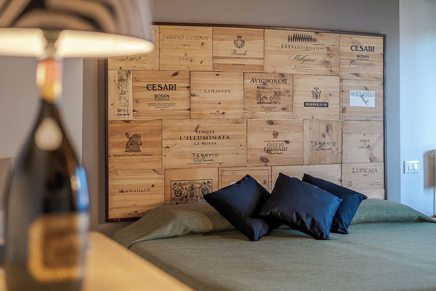 A bedroom with a wooden headboard displaying various wine labels, a bed with dark blue pillows, and a lamp on a bedside table.